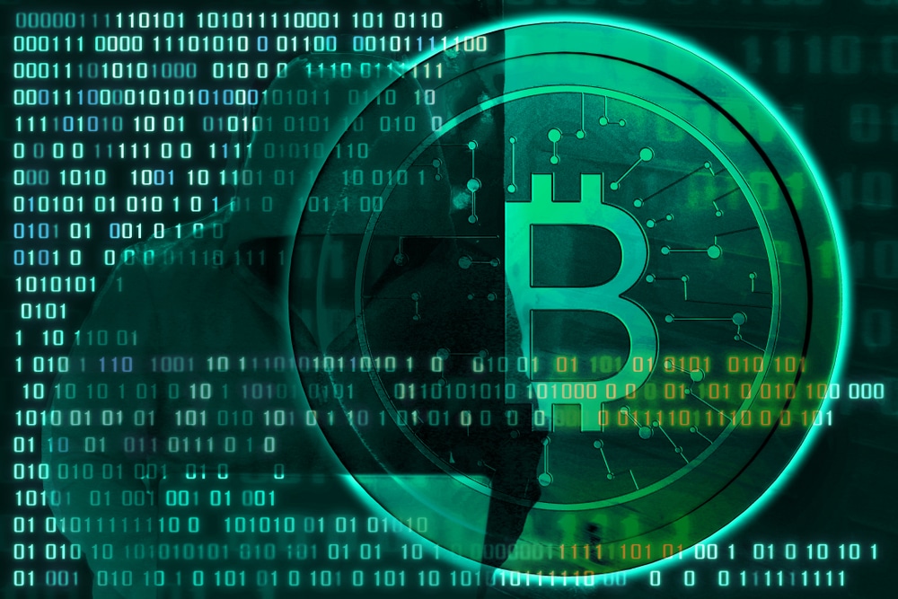Bitcoin next to lines of binary code with a hooded figure on a laptop